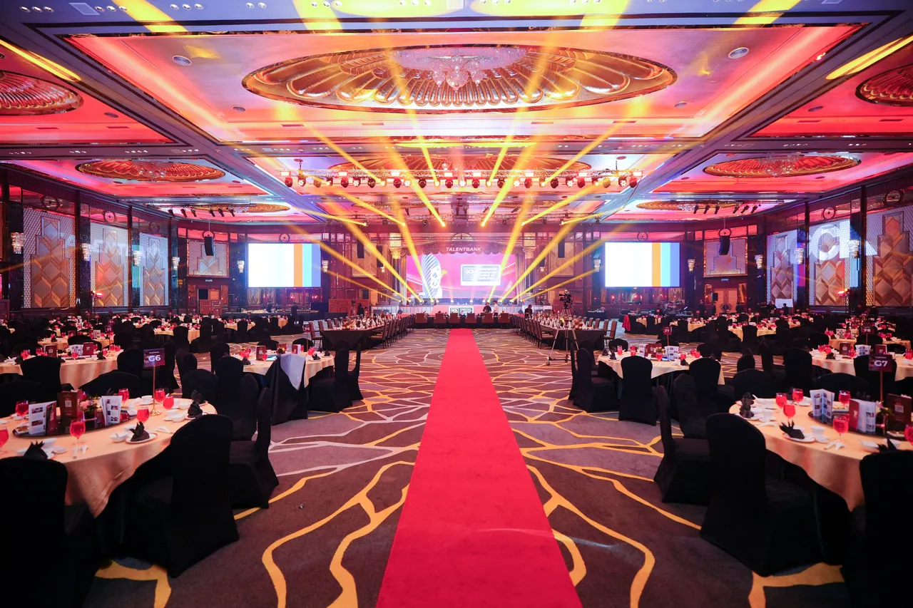 A ballroom set for a Graduates' Choice Award gala, with round tables either side of a red carpet and light beams over the stage