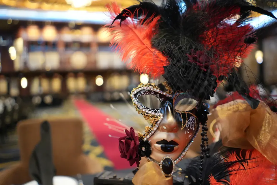A feathered Venetian mask in the foreground, with a ballroom and red carpet behind it