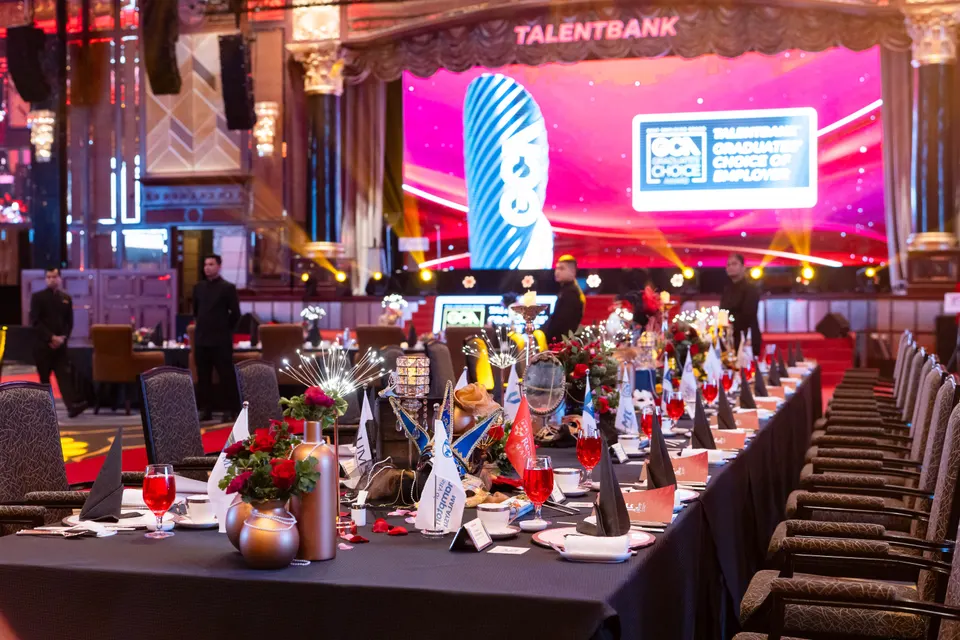 A long gala table dressed with flowers and flags, with the stage and screen behind it
