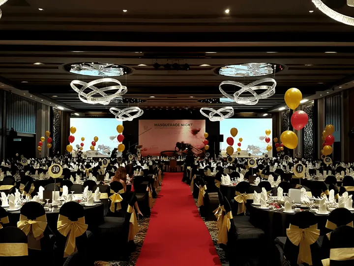 A ballroom set for an annual dinner, with a red carpet aisle, black and gold chair covers and balloons