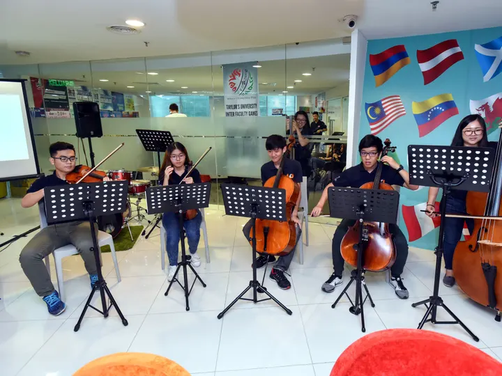 A string ensemble playing in a bright hall with a wall of flags, at a hi-tea