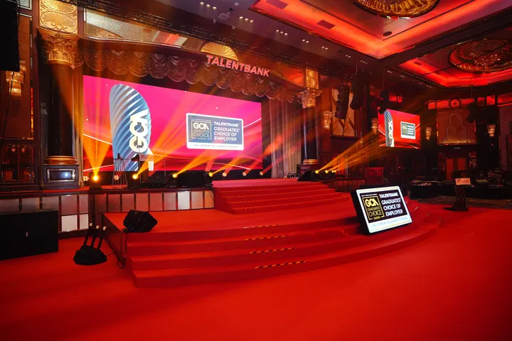A red stage with steps, a large screen and light beams, set for an awards gala