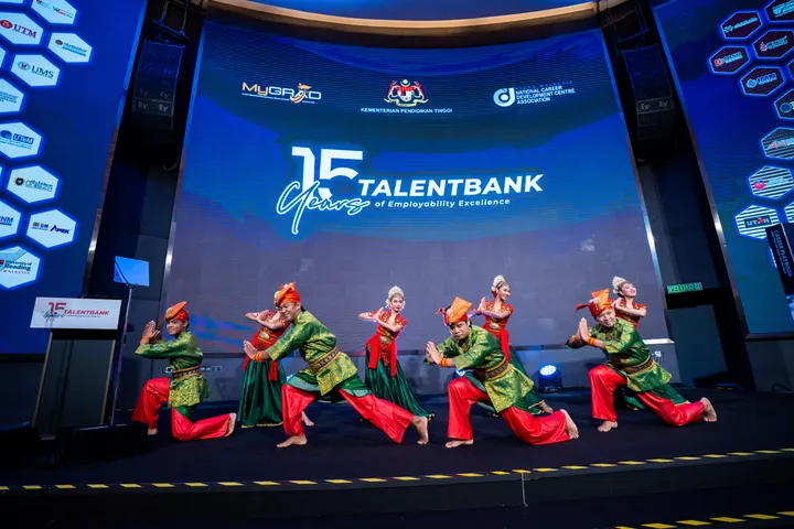 Traditional dancers in green and red costumes on a stage in front of a large LED screen