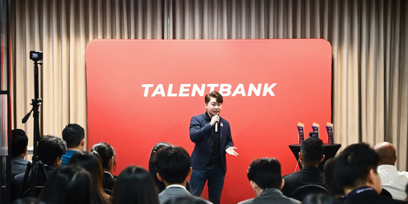 A host with a microphone addressing a seated audience in front of a red Talentbank backdrop