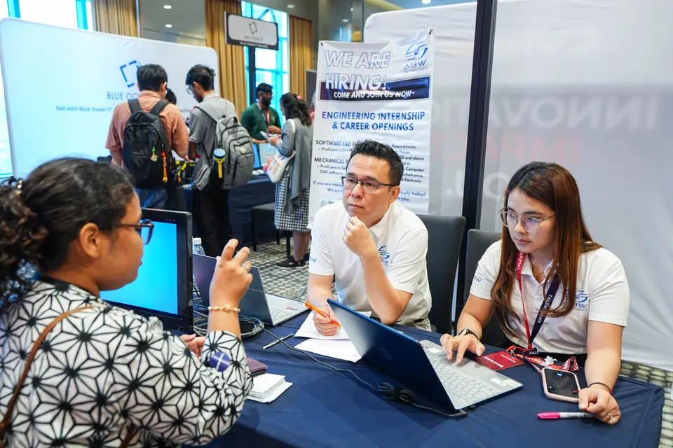 Two employer representatives talking with a candidate at their booth at a Talentbank tech career fair, beneath a We are hiring banner