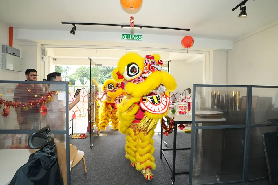Two lion dancers coming through the glass entrance of the Talentbank office, with open-plan desks and ceiling lights behind them