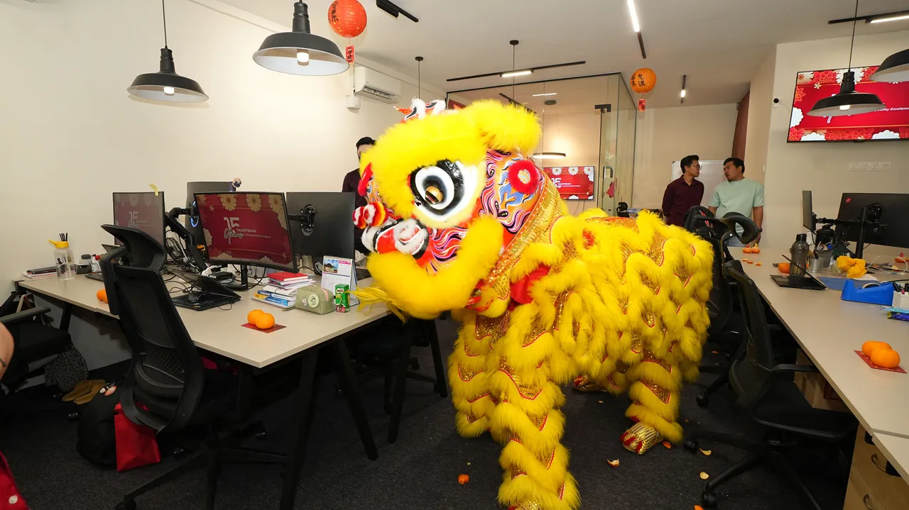 Desks, monitors and pendant lights in the open-plan Talentbank office, with a lion dancer passing between the desks during Chinese New Year 2025