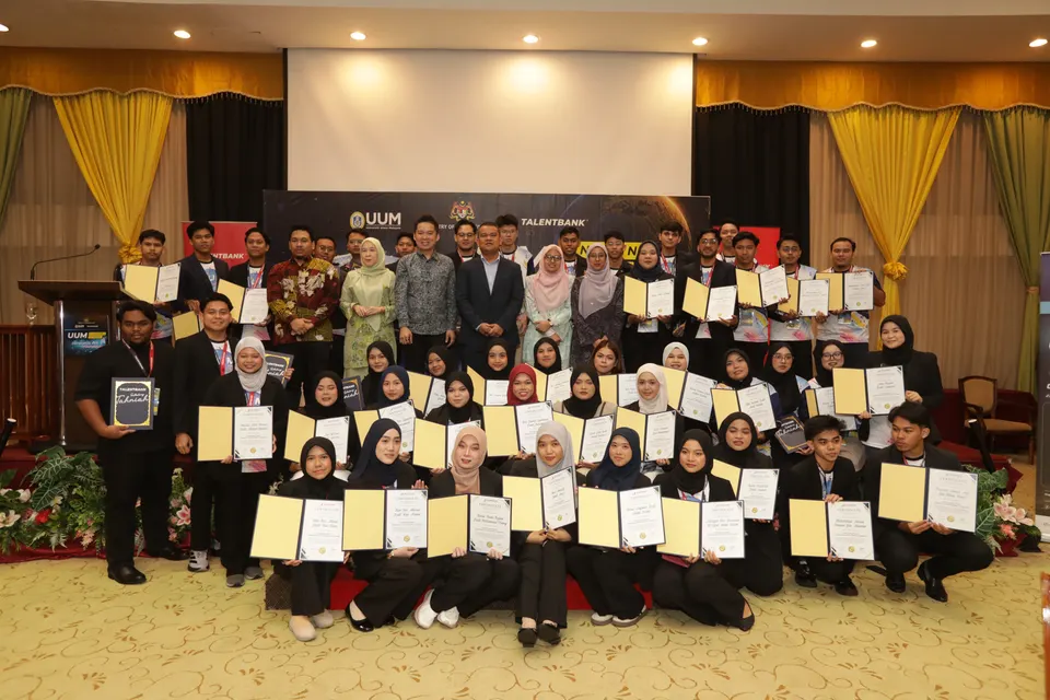 A large group of graduates in formal dress holding their certificates on stage with guests, on graduation day
