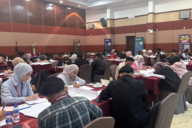A hall of learners seated at tables, writing and working on shared sheets during a Finishing School session