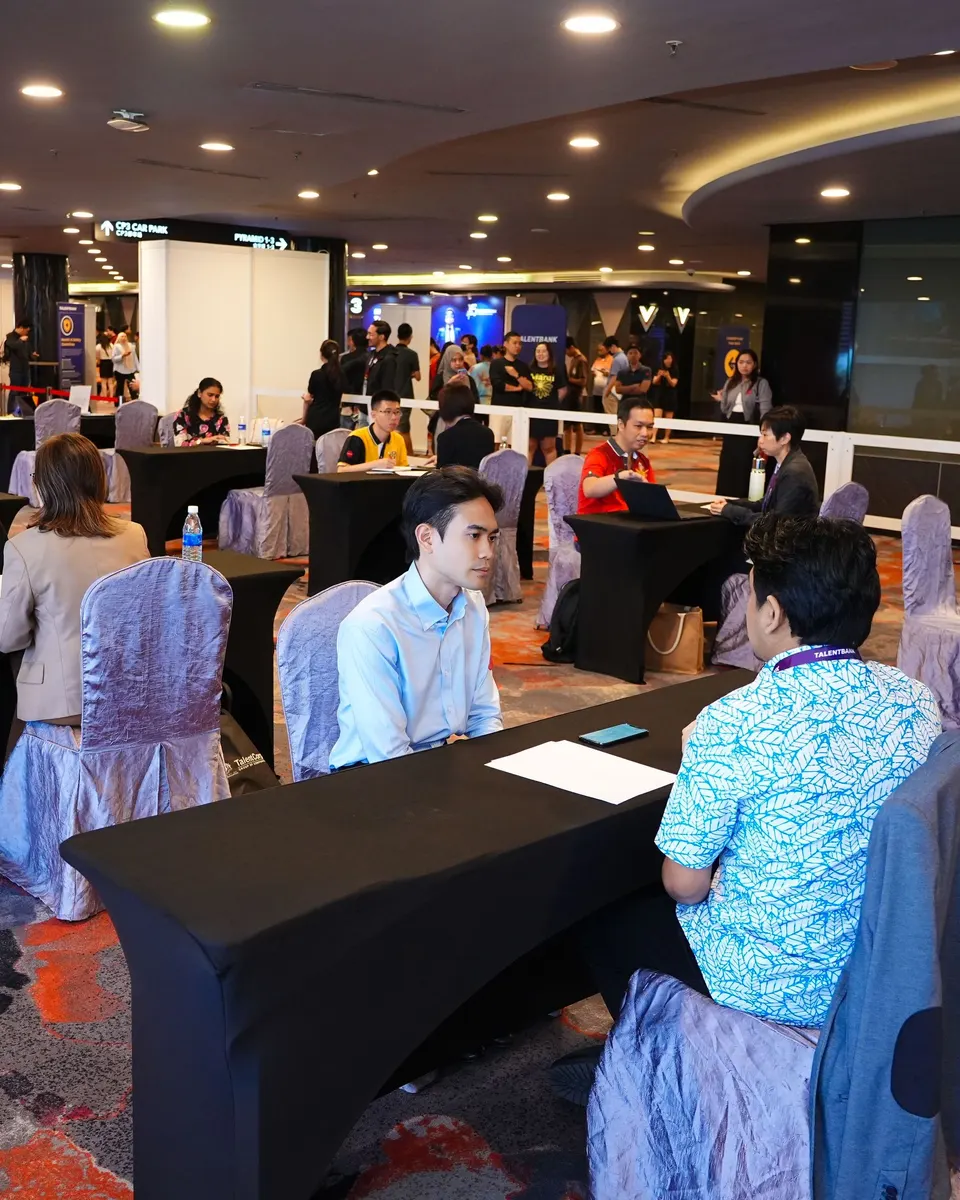 A candidate in a one-to-one interview across a table at the Talentbank Career Fair, with other interviews under way behind