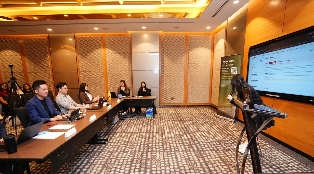 A presenter at a large screen while the judging panel sits at a long table in the Grand Finale room at the 2026 Tech Hackathon
